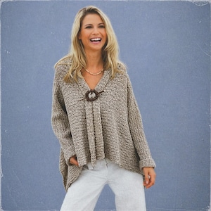 May include: A beige knit sweater with a V-neck and a decorative brown clasp. The sweater has long sleeves and a loose fit. The person is wearing light gray pants and a beaded necklace. The background is a solid light blue.