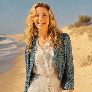 May include: A blue crochet bolero jacket with a scalloped edge. The open-front jacket is worn over a white blouse and dress. The background shows a beach with sand and ocean.