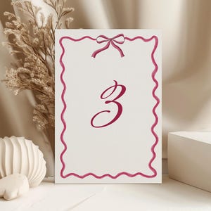 Op de afbeelding: Een witte tafelnummerkaart met een magenta geschulpte rand en strik aan de bovenkant. Het nummer "3" is geschreven in een magenta scriptlettertype. De kaart wordt weergegeven op een wit oppervlak.