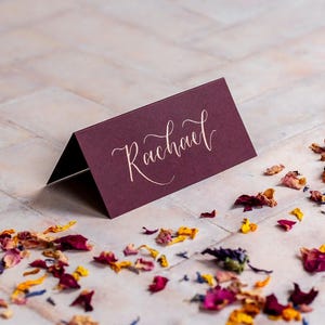Puede incluir: Una tarjeta de mesa burdeos con el nombre "Rachael" en caligrafía elegante. La tarjeta está doblada y de pie. Pétalos de flores secas en tonos rojos, amarillos y morados están esparcidos alrededor de la tarjeta.