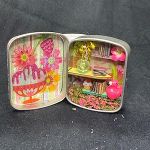 May include: A miniature diorama inside a small, open tin. One side features a dessert with flowers, the other a bookshelf, flamingo, and flowers. The diorama is colorful with pink, yellow, and green accents.