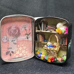 May include: A miniature Easter-themed diorama inside a small, open tin. The scene includes a bookshelf with tiny books, a basket of Easter eggs, a white bunny, and colorful flowers. The tin's interior features a floral design.