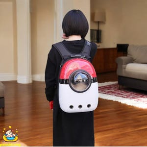 Puede incluir: Una mochila transportadora para mascotas roja, blanca y negra con una ventana redonda y transparente. La mochila tiene una parte superior redondeada y tres orificios de ventilación. La mochila la lleva una persona con un atuendo negro.