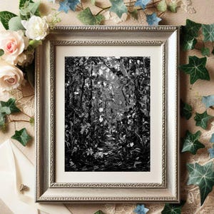 May include: A framed black and white painting of a forest scene. The artwork is in a silver-colored frame, surrounded by floral and leaf decorations. The painting depicts a path through a forest with light filtering through the trees.