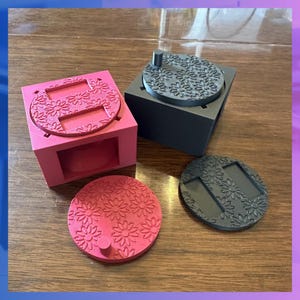 Pode incluir: Uma coleção de caixas e tampas impressas em 3D. Uma caixa e tampa rosa, e uma caixa e tampa cinza, cada uma com um padrão floral. Duas tampas adicionais também são visíveis, uma rosa e uma cinza.