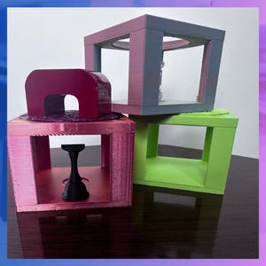 K&ouml;nnte beinhalten: Ein Stapel von drei farbenfrohen, geometrischen, 3D-gedruckten Objekten. Die Objekte sind w&uuml;rfelf&ouml;rmig mit offenen Zentren. Das oberste Objekt ist grau und rosa, das mittlere ist rosa und das untere ist gr&uuml;n. Ein schwarzes, sanduhrf&ouml;rmiges Objekt befindet sich im rosa W&uuml;rfel.