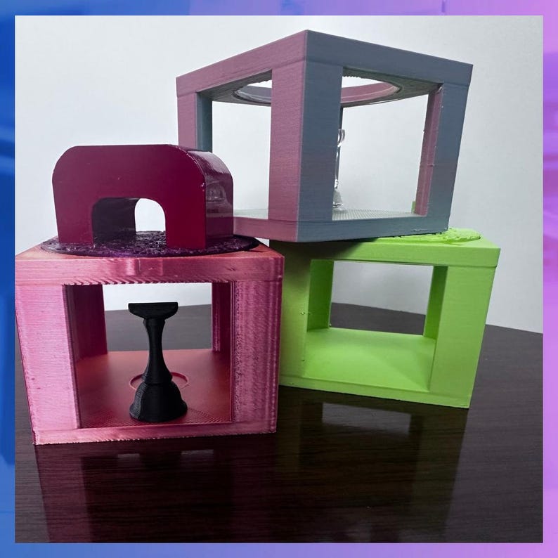 K&ouml;nnte beinhalten: Ein Stapel von drei farbenfrohen, geometrischen, 3D-gedruckten Objekten. Die Objekte sind w&uuml;rfelf&ouml;rmig mit offenen Zentren. Das oberste Objekt ist grau und rosa, das mittlere ist rosa und das untere ist gr&uuml;n. Ein schwarzes, sanduhrf&ouml;rmiges Objekt befindet sich im rosa W&uuml;rfel.