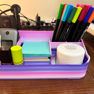 Op de afbeelding: Een bureau-organizer met een verloopontwerp in tinten paars, roze en wit. Het bevat diverse items, waaronder pennen, plaknotities, een tube lotion en andere kleine containers. De organizer staat op een houten oppervlak.