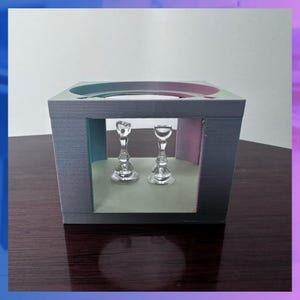 K&ouml;nnte beinhalten: Zwei durchsichtige Kerzenhalter aus Glas mit verzierten Stielen und runden Basen werden in einer grauen, kastenf&ouml;rmigen Struktur ausgestellt. Die Struktur hat eine rechteckige &Ouml;ffnung und einen hellgr&uuml;nen Innenraum.