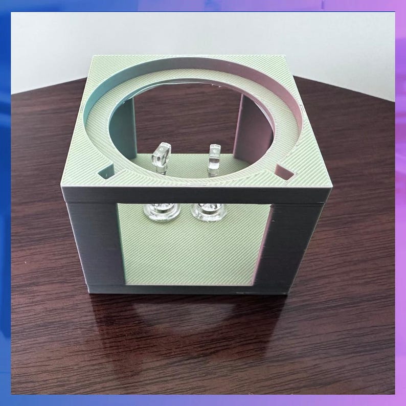 K&ouml;nnte beinhalten: Ein w&uuml;rfelf&ouml;rmiger Pr&auml;sentationsst&auml;nder mit einer kreisf&ouml;rmigen &Ouml;ffnung oben. Der St&auml;nder ist hellgr&uuml;n mit schwarzen Akzenten und enth&auml;lt zwei transparente, dekorative Objekte. Er dient zur Pr&auml;sentation kleiner Gegenst&auml;nde.