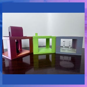 K&ouml;nnte beinhalten: Drei Holz-Pr&auml;sentationsboxen in den Farben Burgunderrot, Gr&uuml;n und Grau. Die bordeauxfarbene Box hat eine gew&ouml;lbte Oberseite, die gr&uuml;ne Box hat einen kleinen Pfosten und die graue Box enth&auml;lt zwei Glasobjekte. Die Boxen stehen auf einer dunkelbraunen Oberfl&auml;che.