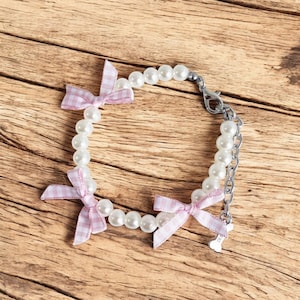 May include: A white pearl bracelet with three pink gingham bows. The bracelet has a silver clasp and a silver charm in the shape of a bone.