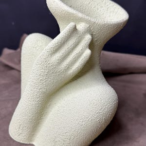 Pode incluir: Um vaso de cerâmica verde claro em forma de figura, com uma superfície texturizada. O vaso apresenta uma forma curva com um braço e uma mão que alcançam a abertura. Um objeto decorativo.