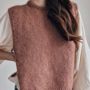 May include: A dusty rose knitted sweater vest featuring a ribbed crew neckline and hem, worn over a cream-colored long-sleeved shirt with voluminous sleeves and paired with blue denim jeans. The vest has a textured knit pattern along the armholes.