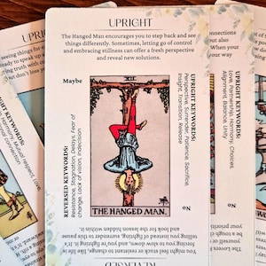 May include: The Hanged Man tarot card features a man hanging upside down from a tree branch. The card is surrounded by text that includes the keywords "Upright" and "Reversed" along with descriptions of the card's meaning.