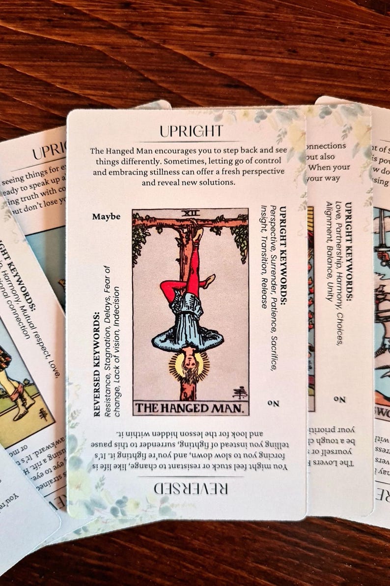 May include: The Hanged Man tarot card features a man hanging upside down from a tree branch. The card is surrounded by text that includes the keywords "Upright" and "Reversed" along with descriptions of the card's meaning.