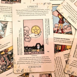 May include: A close-up of a tarot card reading spread. The Ace of Pentacles card is in the center of the spread. The card features a hand holding a gold coin with a five-pointed star in the center. The card is surrounded by other tarot cards, including the Queen of Swords, the Death card, and the 12 of Swords.
