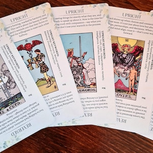 May include: Four tarot cards with colorful illustrations and text. The cards are laid out on a wooden surface. The cards are from the Rider-Waite tarot deck. The cards are: Two of Cups, Two of Swords, Six of Wands, and Two of Lovers.