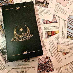 May include: A close-up of a green tarot card with a gold crescent moon and star design. The card is surrounded by other tarot cards with white backgrounds and black text.