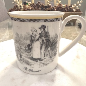 May include: White ceramic mug with a black and white illustration of a couple walking in a forest. The mug has a gold trim and the words "La Rencontre" at the bottom.