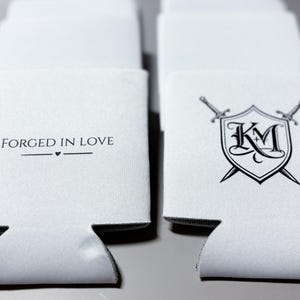 May include: White beverage insulators with black text and graphics. One reads "FORGED IN LOVE" with a heart symbol. Another features a shield with "KM" and crossed swords. These are designed to keep drinks cold.