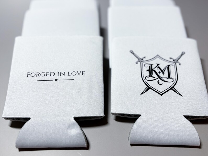 May include: White beverage insulators with black text and graphics. One reads "FORGED IN LOVE" with a heart symbol. Another features a shield with "KM" and crossed swords. These are designed to keep drinks cold.