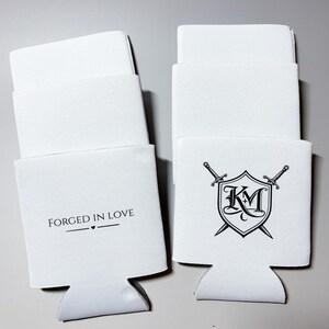 May include: White beverage insulators with a cut-out base. One has the text "FORGED IN LOVE" with a heart symbol. The other features a shield design with the letters "KM" and crossed swords. Ideal for keeping drinks cold.
