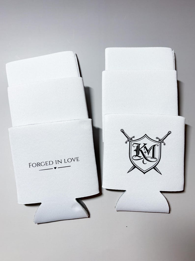 May include: White beverage insulators with a cut-out base. One has the text "FORGED IN LOVE" with a heart symbol. The other features a shield design with the letters "KM" and crossed swords. Ideal for keeping drinks cold.