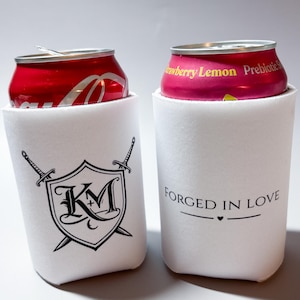 May include: Two white can coolers with black graphics and text. One cooler features a shield design with the letters "KM" and crossed swords. The other cooler has the text "FORGED IN LOVE" with a heart symbol. Cans of soda are inside.