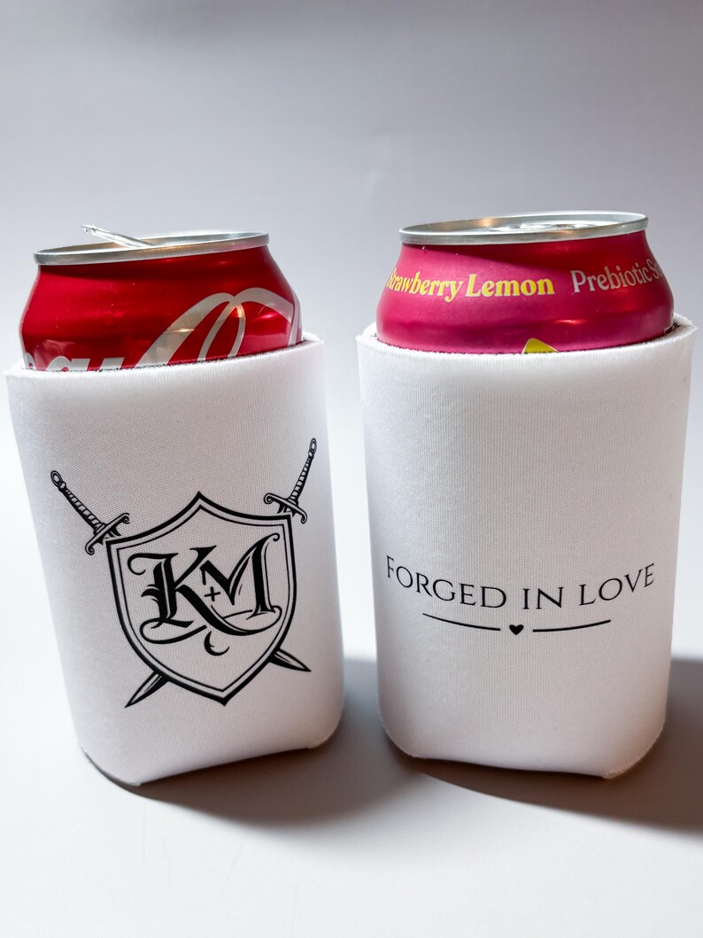 May include: Two white can coolers with black graphics and text. One cooler features a shield design with the letters "KM" and crossed swords. The other cooler has the text "FORGED IN LOVE" with a heart symbol. Cans of soda are inside.