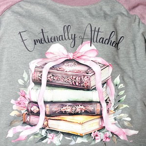 May include: Gray raglan-style t-shirt with a pink neckline and sleeves. The shirt features a stack of books tied with a pink ribbon and floral accents. Text reads "Emotionally Attached to Fictional Characters."