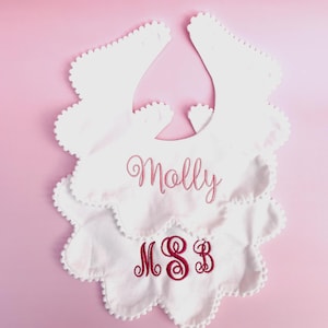May include: Two white baby bibs with scalloped edges and a pink background. One bib has the name "Molly" embroidered in pink script. The other bib has the initials "MSB" embroidered in a burgundy color.