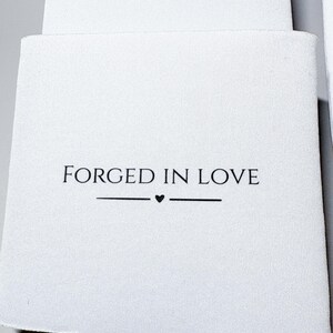 May include: White can cooler with the text "FORGED IN LOVE" in black, with a small heart symbol. The cooler is made of a soft, textured material and is designed to keep beverages cold. The design is simple and elegant.
