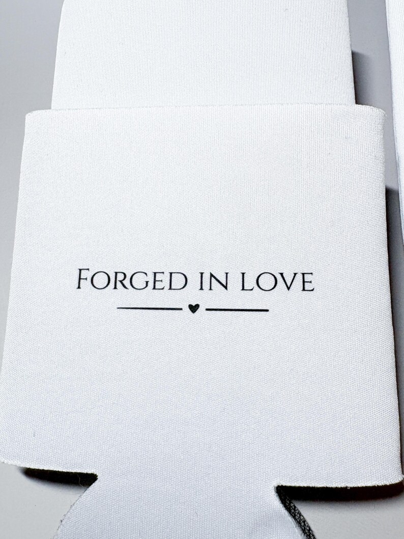 May include: White can cooler with the text "FORGED IN LOVE" in black, with a small heart symbol. The cooler is made of a soft, textured material and is designed to keep beverages cold. The design is simple and elegant.
