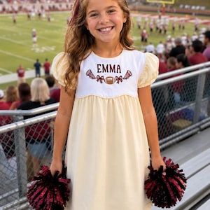 May include: A cream-coloured dress with puff sleeves and a white bodice featuring the embroidered name EMMA in maroon, flanked by two maroon megaphones, two maroon bows, and a football. The dress is paired with two maroon and black pom-poms.