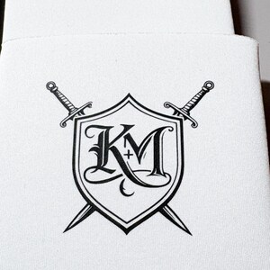 May include: White beverage insulator featuring a black shield design with the letters "KM" and a cross, crossed by two swords. The design is centered on the insulator, with a simple, clean aesthetic.