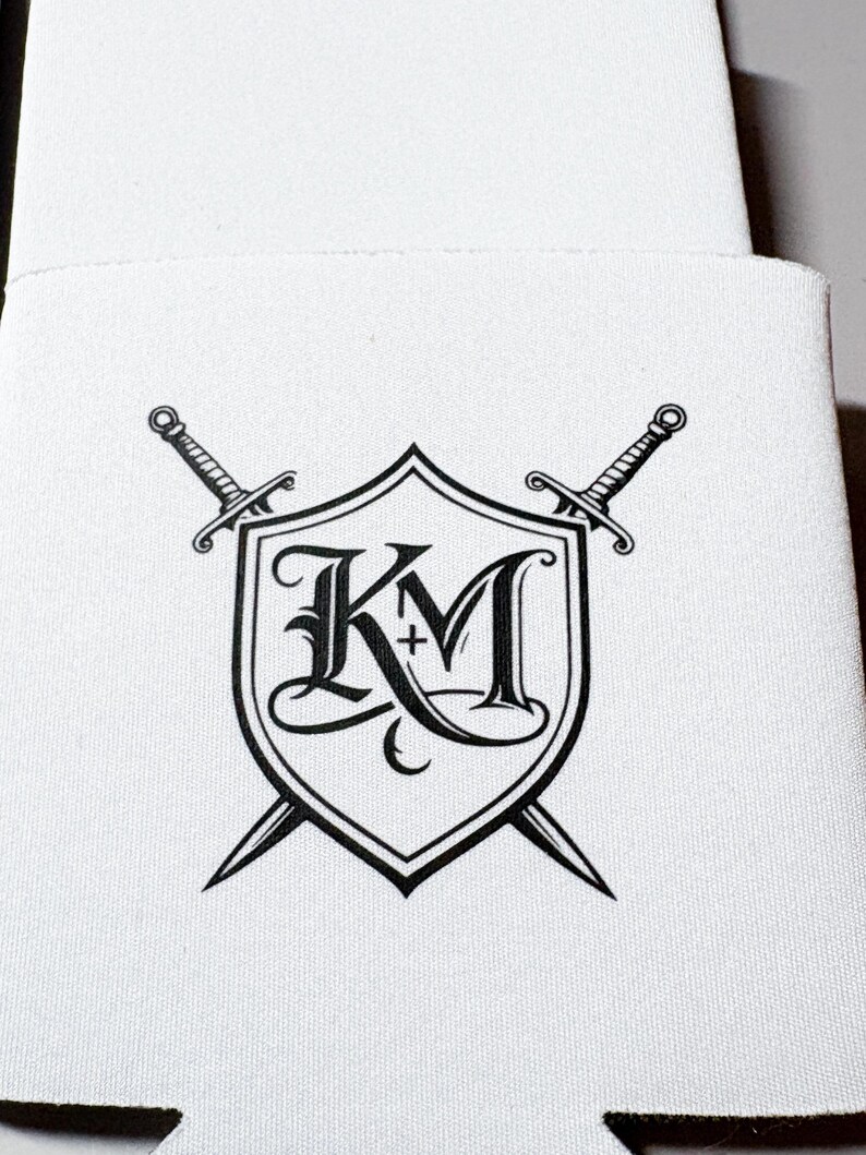 May include: White beverage insulator featuring a black shield design with the letters "KM" and a cross, crossed by two swords. The design is centered on the insulator, with a simple, clean aesthetic.