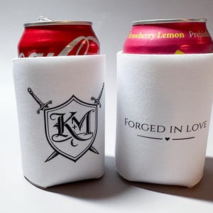 May include: Two white can coolers. One cooler has a black shield design with crossed swords and the letters "KM+". The other cooler has the text "FORGED IN LOVE" with a heart symbol. Each cooler holds a soda can.