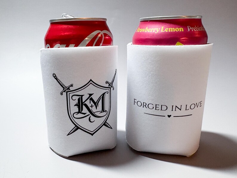 May include: Two white can coolers. One cooler has a black shield design with crossed swords and the letters "KM+". The other cooler has the text "FORGED IN LOVE" with a heart symbol. Each cooler holds a soda can.