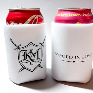 May include: Two white can coolers with black text and graphics. One cooler has a shield design with the letters "KM" and crossed swords. The other cooler says "Forged in Love" with a heart symbol. Each cooler holds a red can.