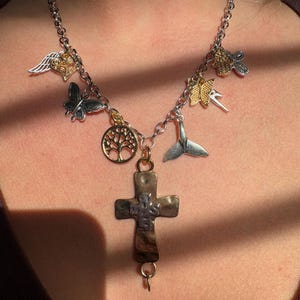 May include: A silver chain necklace with multiple charms, including a cross, a tree, a butterfly, a heart with wings, a fish, and a bird. The charms are gold and silver.