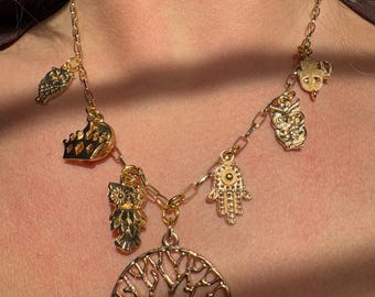 May include: A gold chain necklace with multiple charms, including a tree of life, an owl, a hamsa hand, and a crown. The charms are all gold-toned and have intricate details.