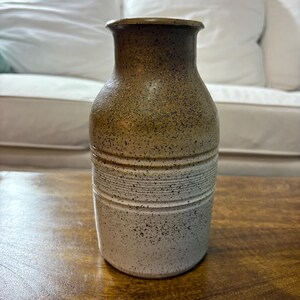 May include: A speckled ceramic vase with a two-tone design. The top portion is a warm brown, transitioning to a speckled white base. Horizontal lines encircle the middle section. The vase is displayed on a wooden surface.