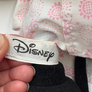 May include: A close-up of a Disney plush toy. The toy has a black head and a white tag with the Disney logo. The toy is wearing a white outfit with a pink floral pattern.