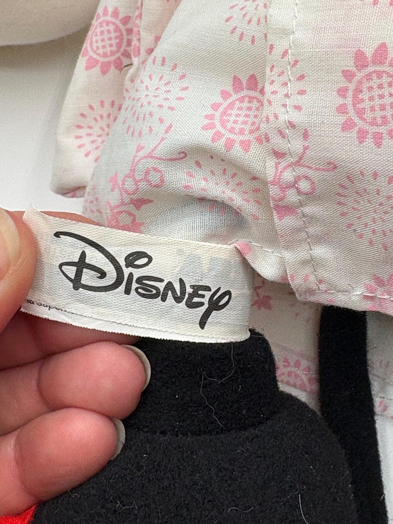 May include: A close-up of a Disney plush toy. The toy has a black head and a white tag with the Disney logo. The toy is wearing a white outfit with a pink floral pattern.