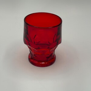 May include: A small, ruby red glass with a textured, faceted design. The shot glass has a cylindrical shape with a wide opening and a detailed base. The glass is approximately 6.35 cm tall.