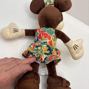 May include: A plush Minnie Mouse doll with brown ears, a matching bow, and a colorful floral dress. The doll has white gloves with black stitched details and brown shoes. The doll is positioned with its arms outstretched.