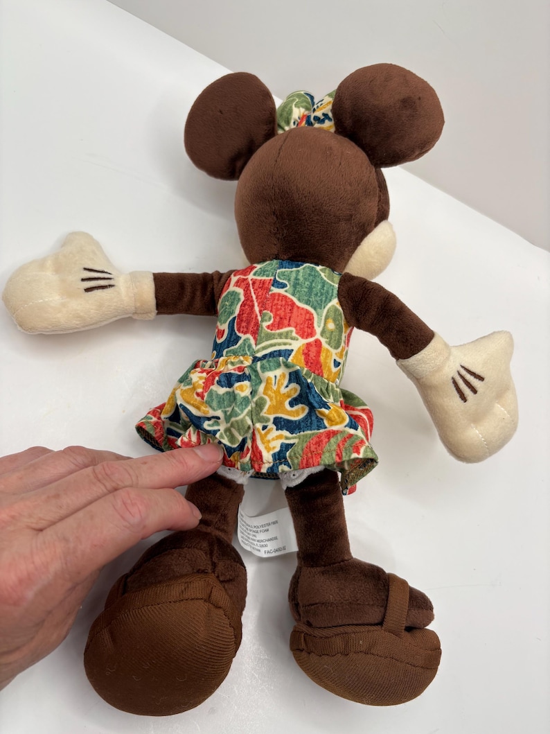 May include: A plush Minnie Mouse doll with brown ears, a matching bow, and a colorful floral dress. The doll has white gloves with black stitched details and brown shoes. The doll is positioned with its arms outstretched.