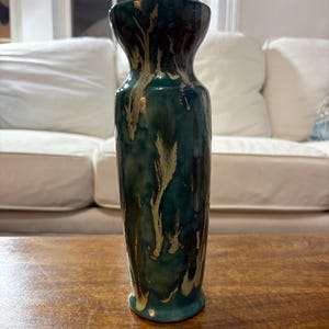 May include: A tall ceramic vase with a flared top and a narrow base. The vase is glazed in shades of teal and dark green, with streaks of beige. The vase is approximately 30 cm tall.