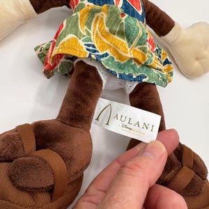 May include: A plush doll wearing a colorful floral print dress and brown sandals. The doll has brown arms and legs, with white gloves and a white face. A tag reads "Aulani, A Disney Resort & Spa".